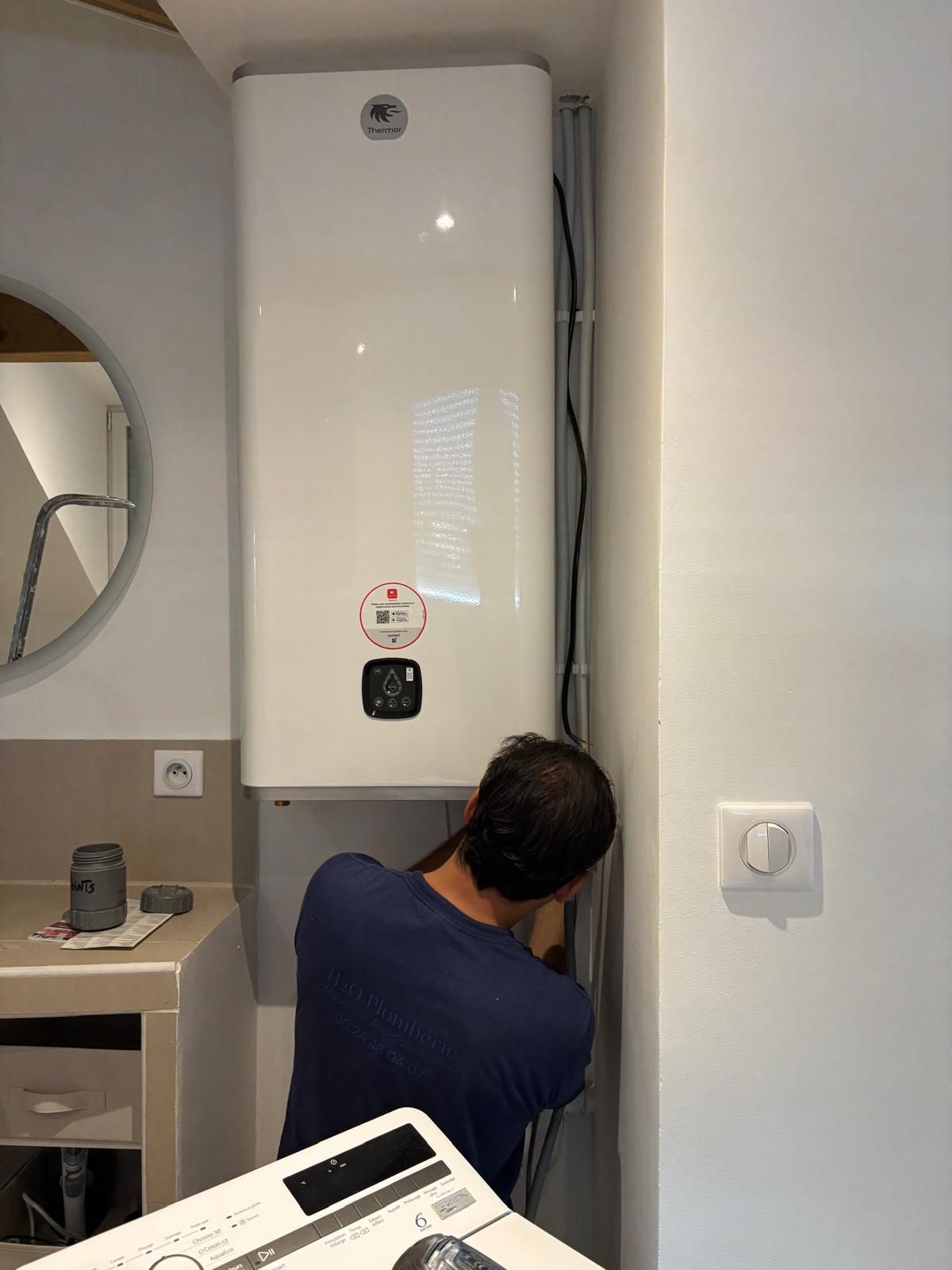 Nicolas, de dos en t-shirt H2O Plomberie, en train de raccorder un chauffe-eau électrique Thermor fixé au mur